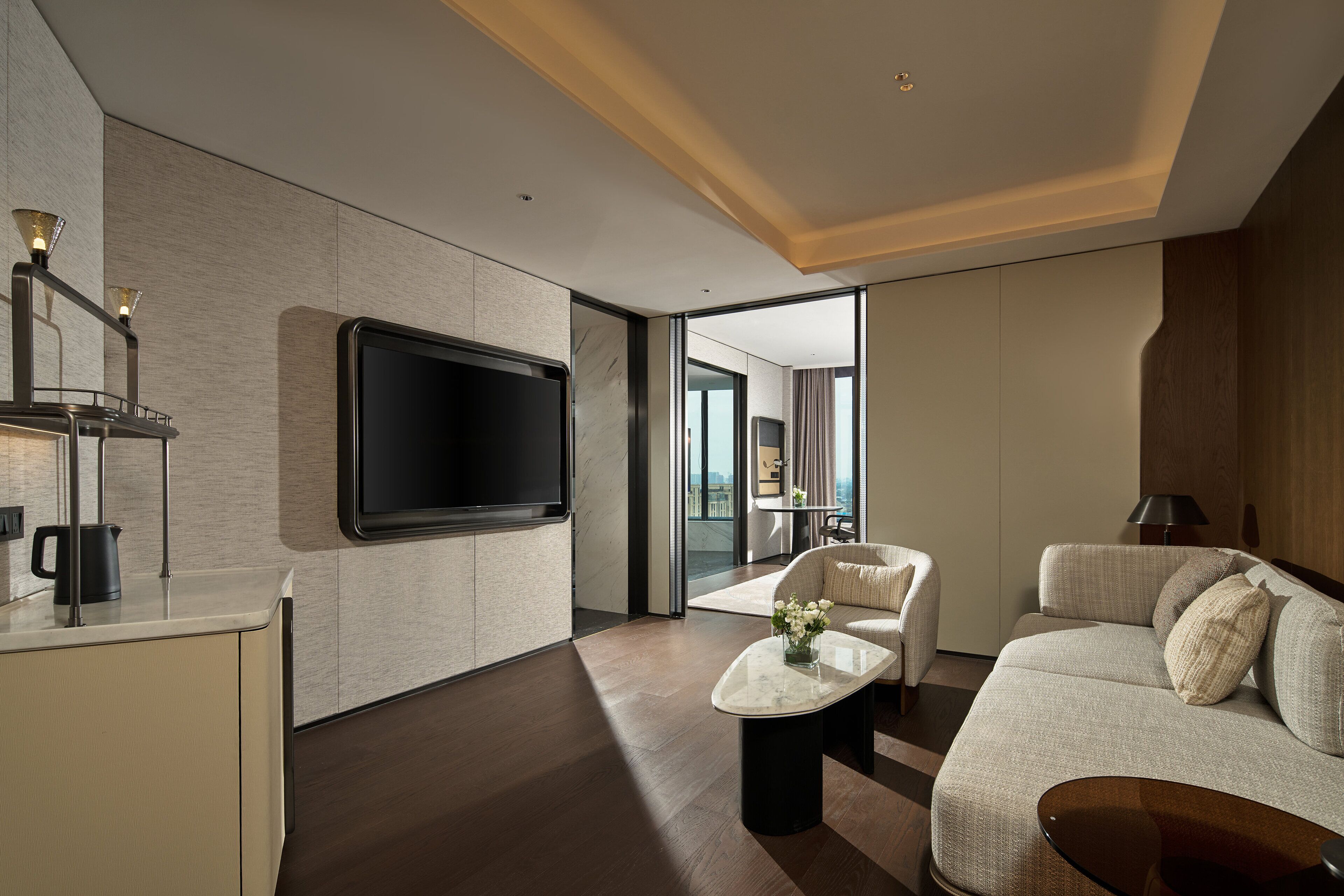 A modern hotel room with a large flat-screen TV, a sofa, a coffee table, and a view of the city through large windows.