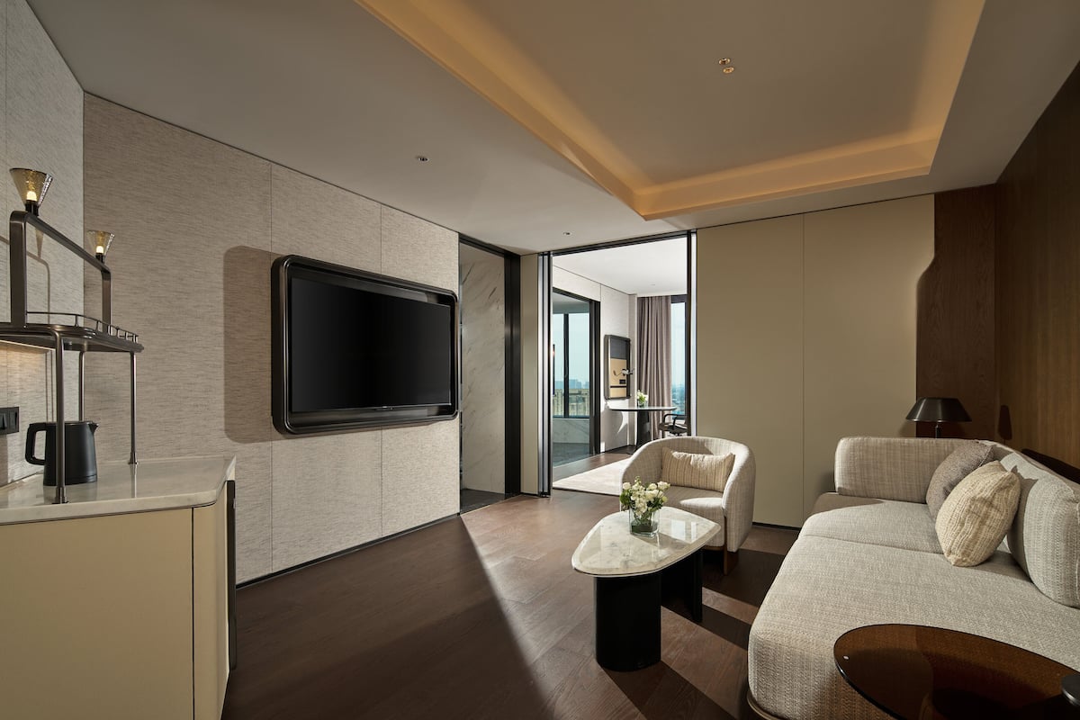 A modern hotel room with a large flat-screen TV, a sofa, a coffee table, and a view of the city through large windows.