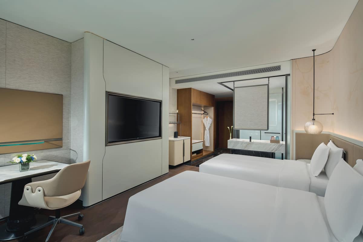 A modern hotel room with a flat-screen TV, a desk with a chair, two beds, a bathroom with a mirror, and a nightstand.