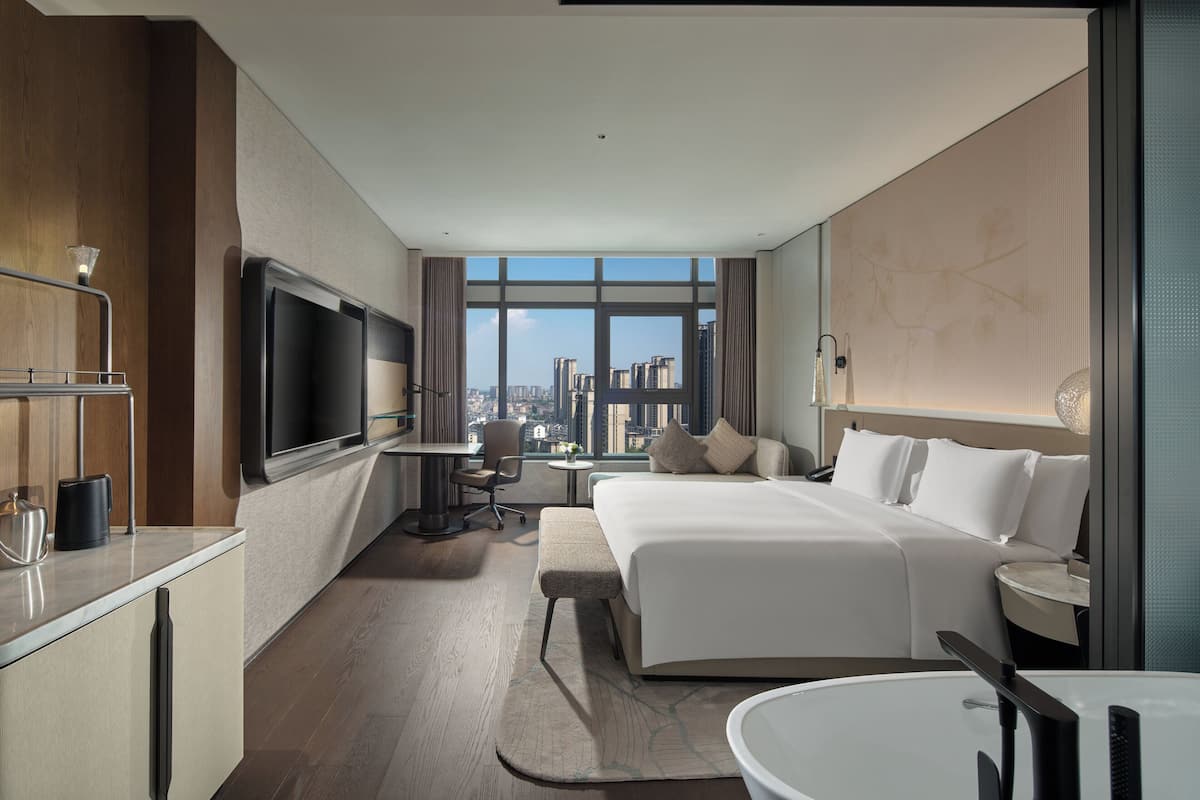A modern hotel room with a large bed, a desk, a TV, and a view of the city.