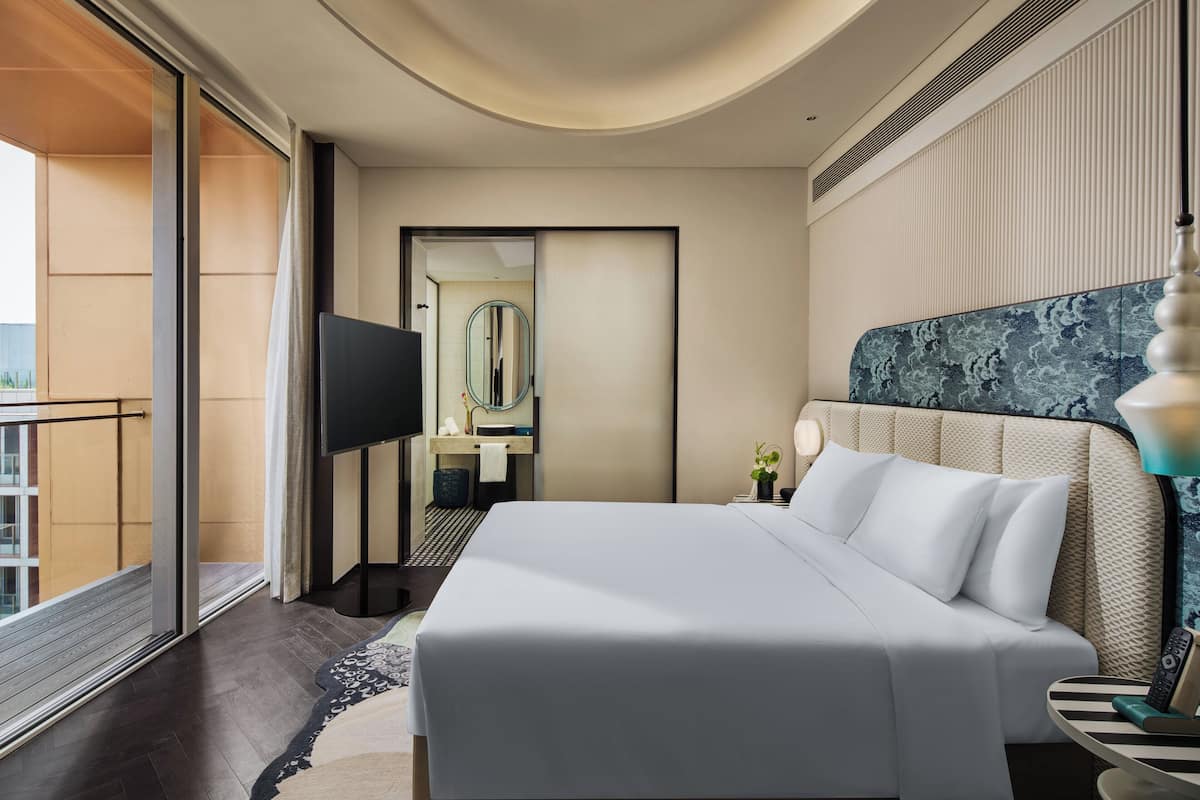 A modern hotel room with a large bed, a television, a bathroom with a mirror, and a balcony.
