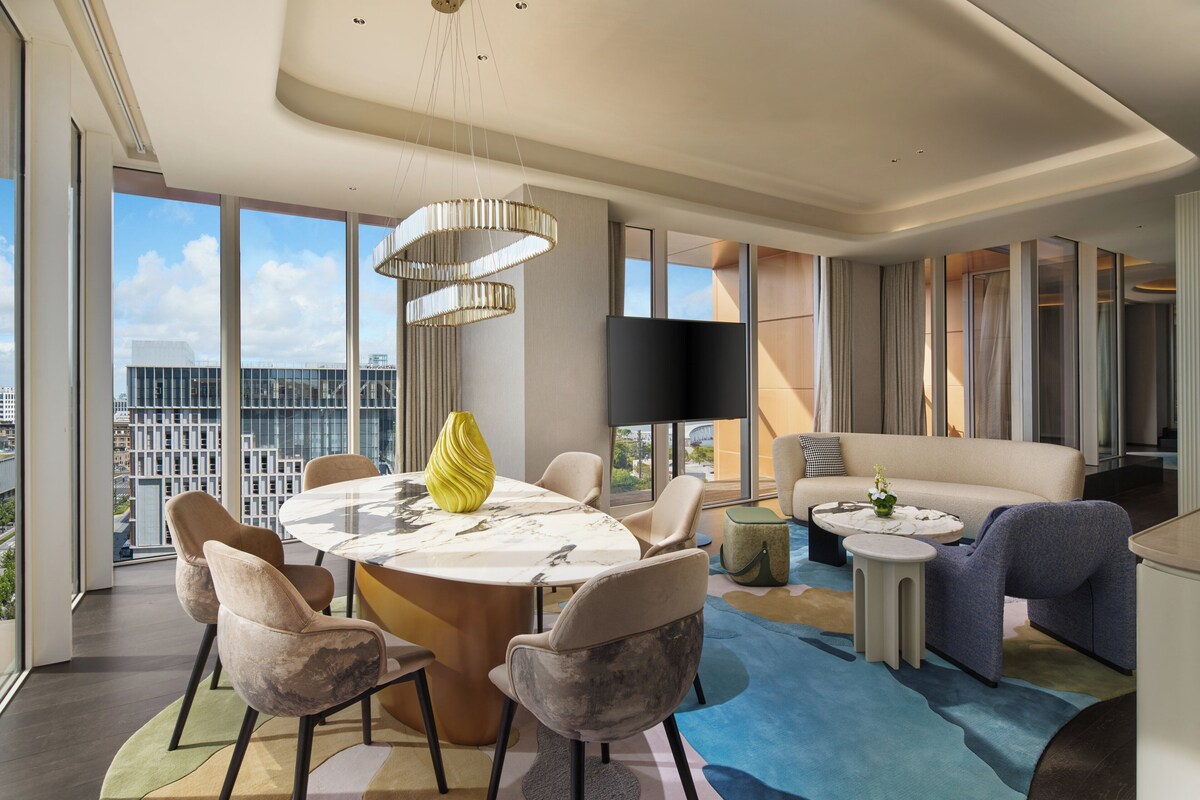 A modern living room with a round dining table, comfortable seating, a flat-screen TV, and a large window offering a city view.