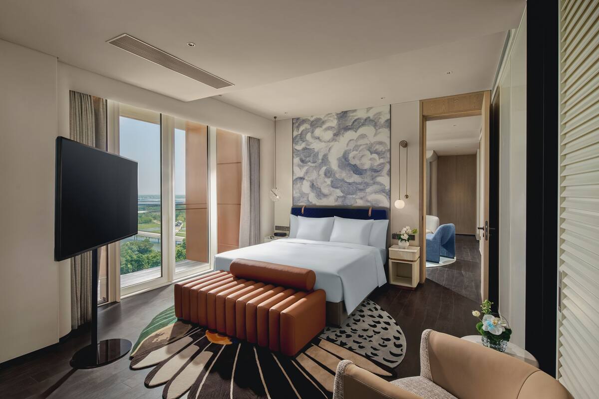 A modern hotel room with a large bed, a flat-screen TV, a view of the city, and a balcony.