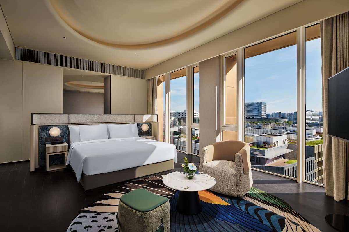 A modern hotel room with a large bed, a sitting area with a chair and a small table, and a balcony with a city view.