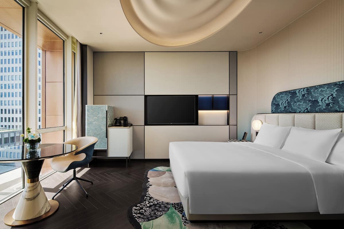 A modern hotel room with a large bed, a desk with a chair, a flat-screen TV, and a view of the city.