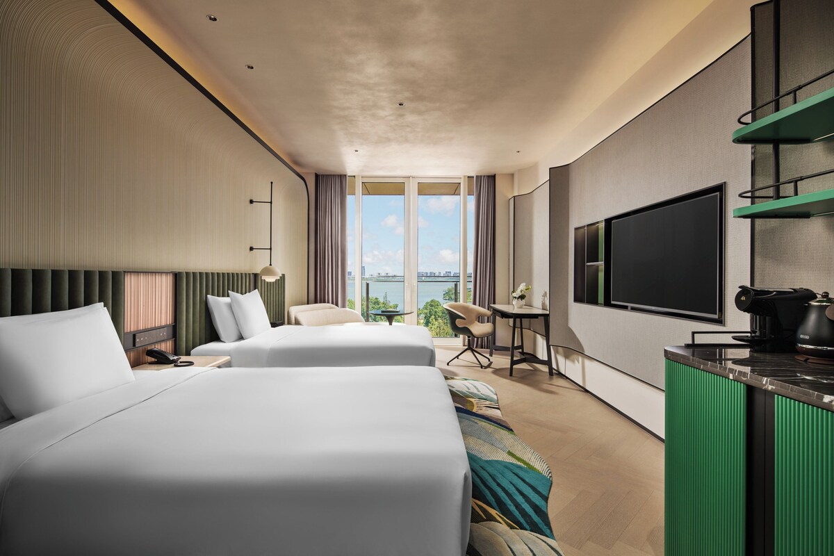 A modern hotel room with two beds, a desk, a chair, a TV, and a view of the city.