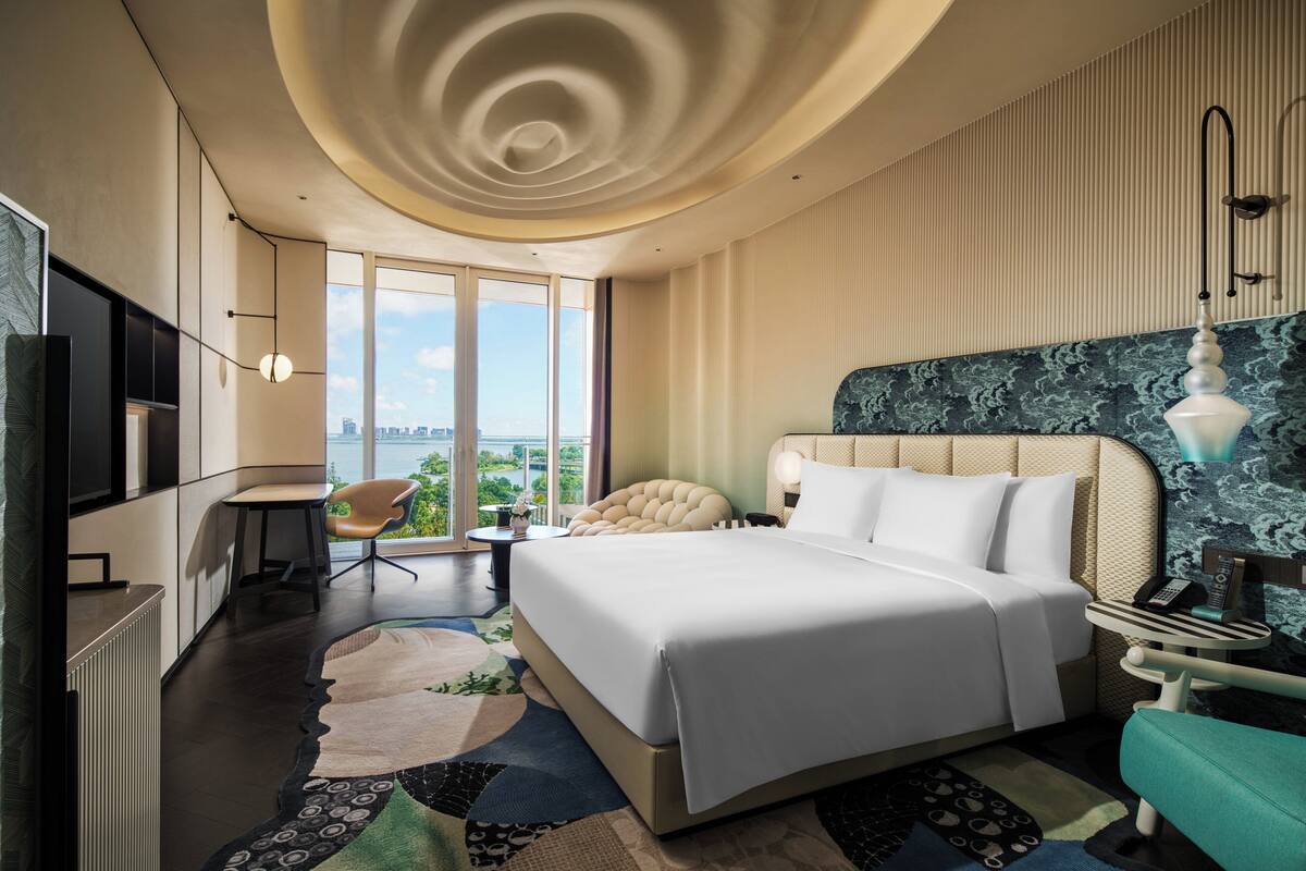 A modern hotel room with a large bed, a desk, a chair, and a view of the city.