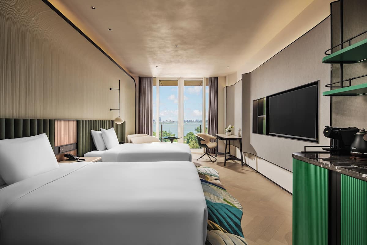 A modern hotel room with two beds, a desk, a chair, a TV, and a view of the city.