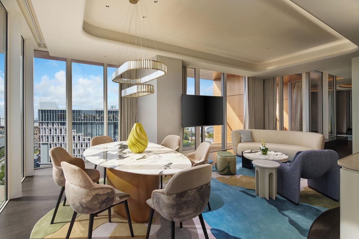 A modern living room with a round dining table, comfortable seating, a flat-screen TV, and a large window offering a city view.