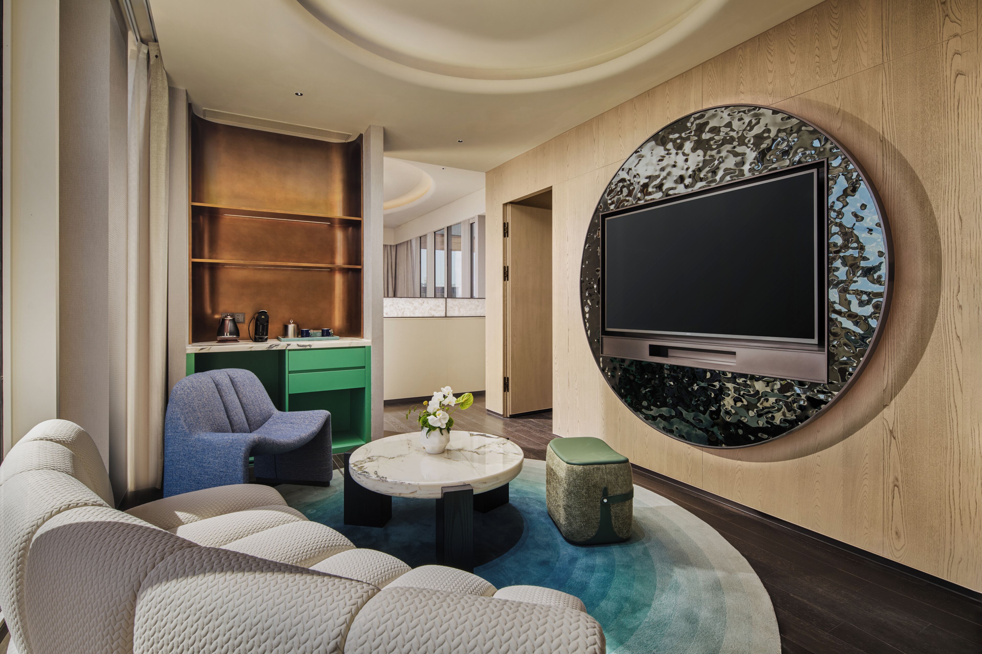 A modern living room with a flat-screen TV mounted on a wooden wall, a round coffee table, and a seating area with sofas and chairs.