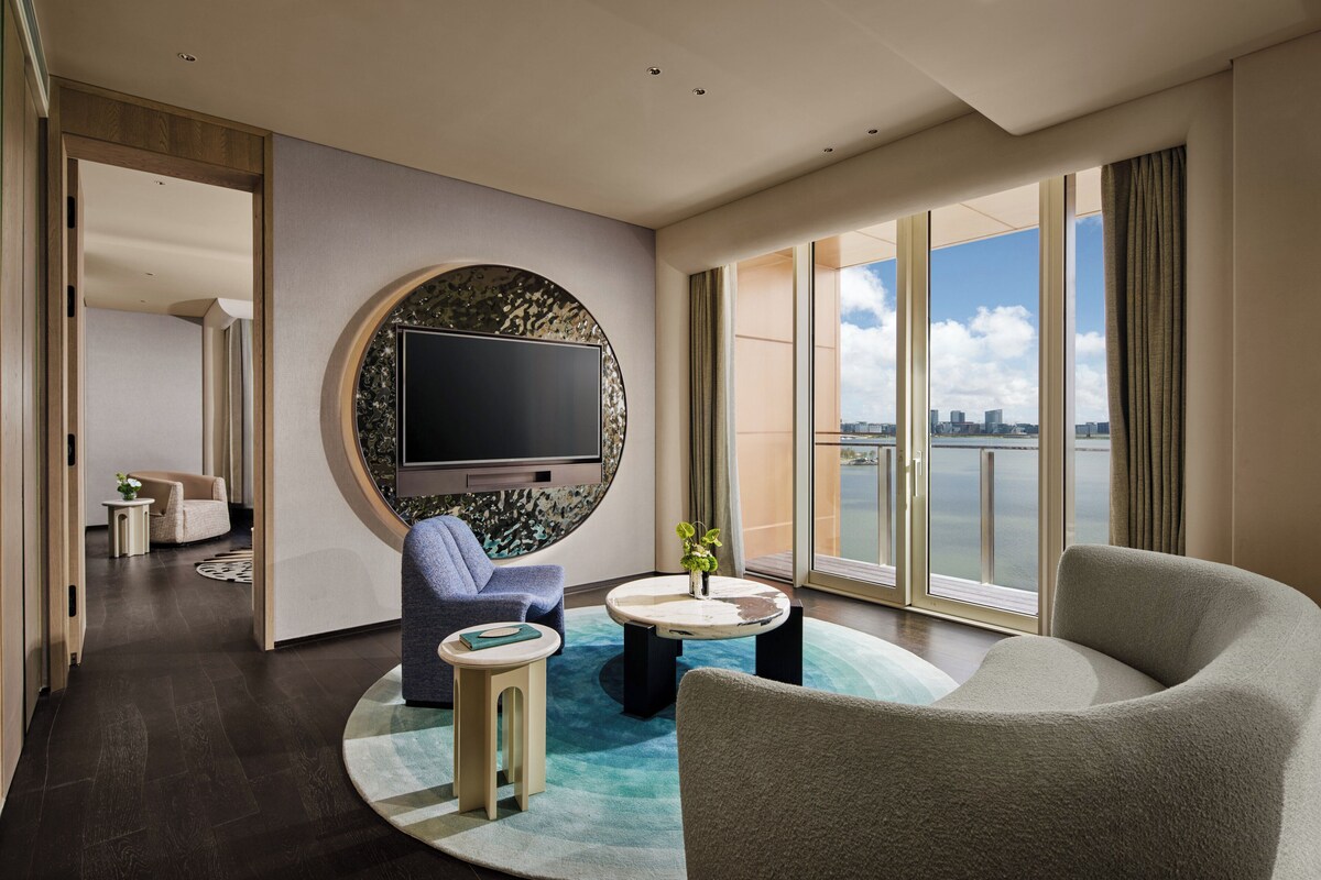 A modern hotel room with a large TV, comfortable seating, a round coffee table, and a view of the city.