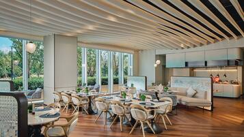 Un espace repas moderne avec des tables et des chaises en bois, de grandes fenêtres et un coin cuisine doté d’un bar.