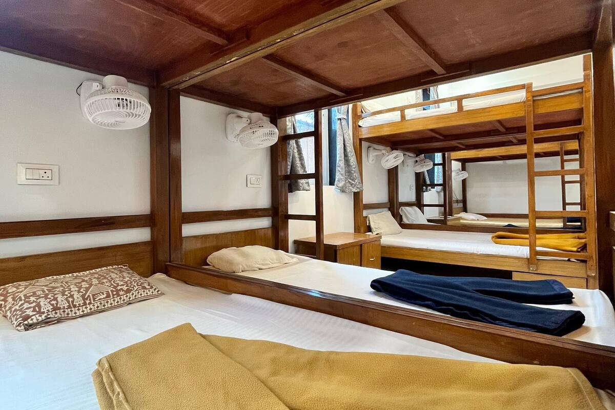 A bunk bed room with a wooden ceiling, a white wall, and a window. There is a bed with a white mattress and a brown blanket.