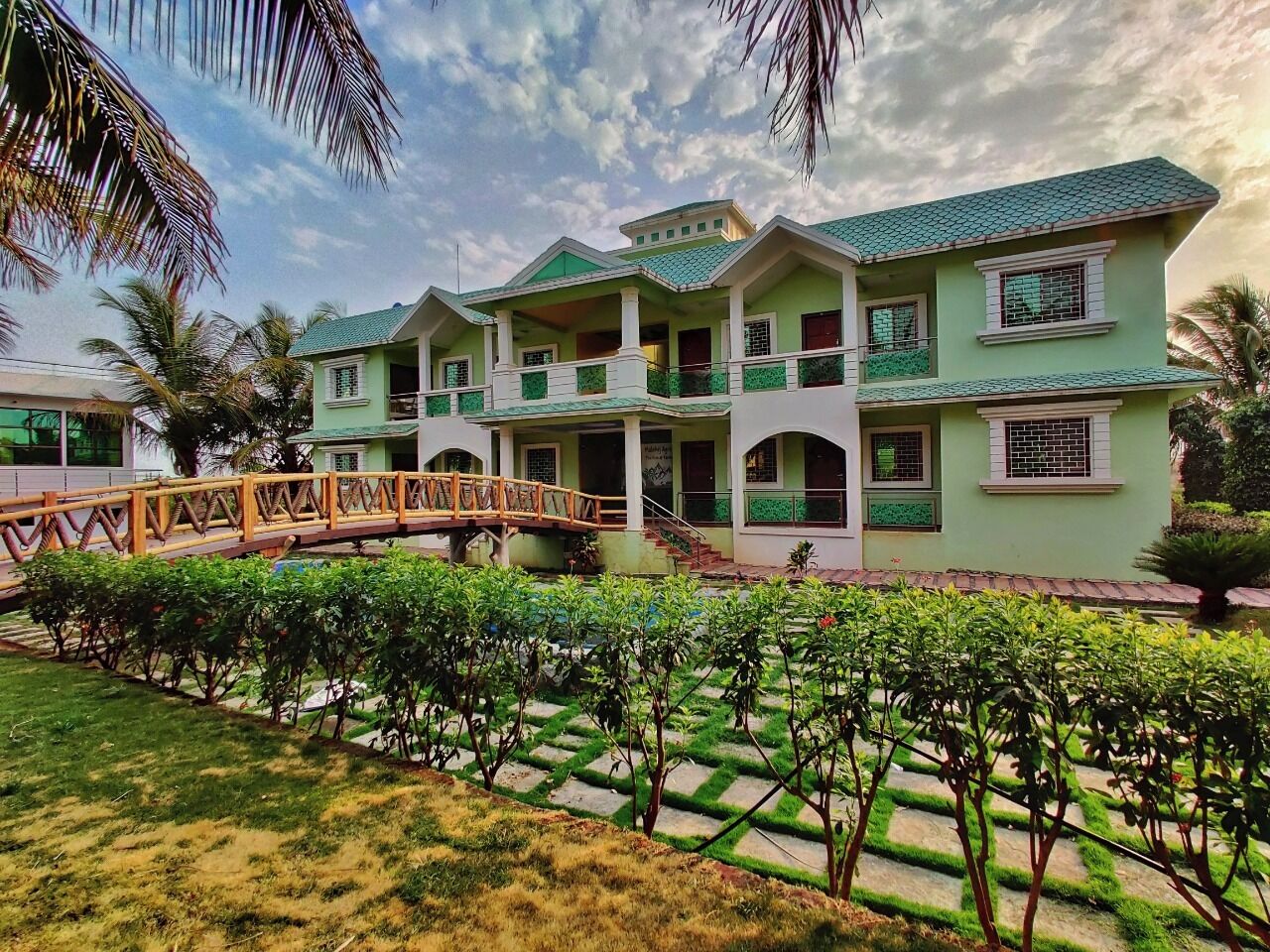 A multi-story building with a green roof and white walls, featuring a central courtyard with a wooden bridge and neatly trimmed hedges.