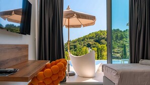 Een moderne hotelkamer met een groot raam, een flatscreen televisie, een houten bureau, een opvallende oranje fauteuil, een witte clubfauteuil en een bed met een beige plaid.