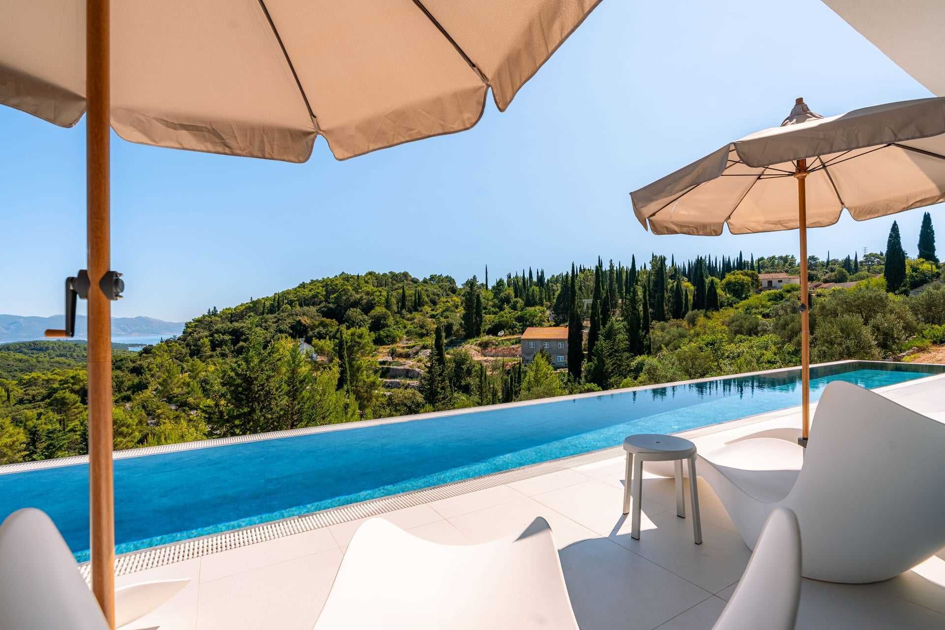 A poolside area with white lounge chairs and beige umbrellas, overlooking a scenic hillside with trees and a clear blue sky.