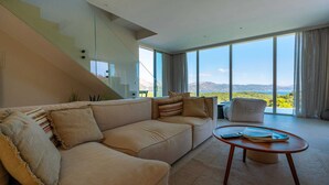 Una sala de estar moderna con un sofá color beige, una mesa de centro redonda y vistas a un paisaje montañoso a través de grandes ventanales.
