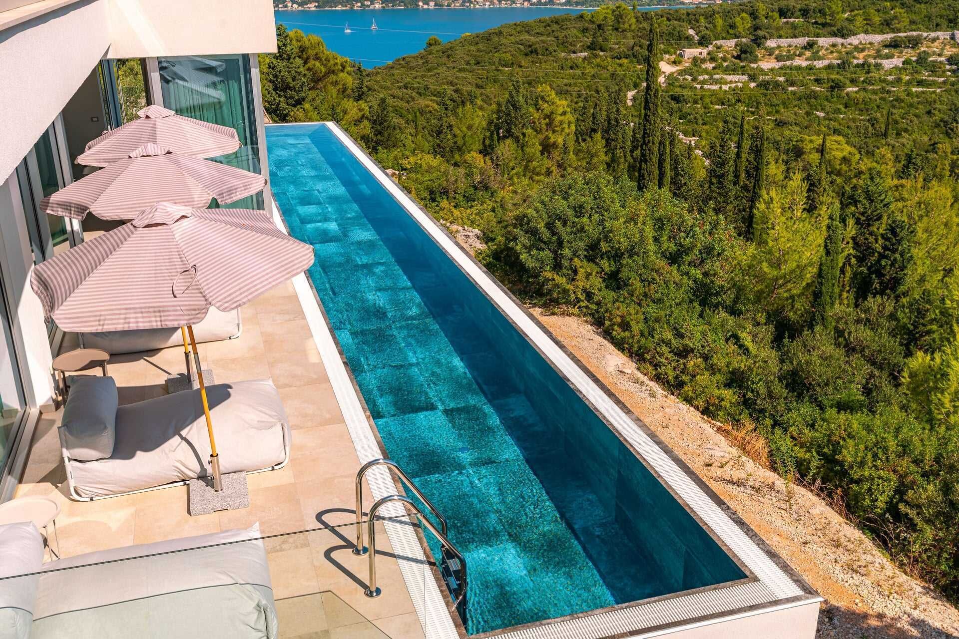 A rooftop infinity pool with lounge chairs and umbrellas, surrounded by lush greenery and a view of the sea.