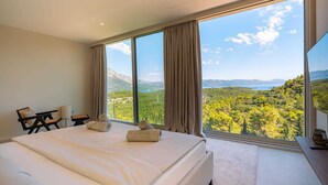 Un dormitorio con una cama grande, un escritorio y una silla, con vistas panorámicas a montañas y árboles a través de amplios ventanales.