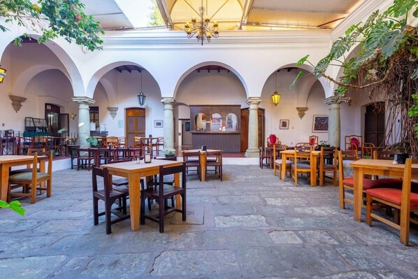 Un comedor interior con varias mesas y sillas de madera, suelo de piedra y arcos.