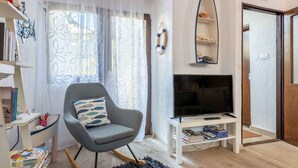 Un salon avec un fauteuil à bascule gris, un téléviseur à écran plat posé sur un meuble blanc, une bibliothèque et un rideau à motifs.
