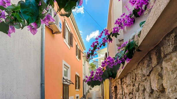 Une rue étroite, bordée de bâtiments des deux côtés, l’un peint en orange et l’autre dans une couleur claire, avec un mur de pierre sur la droite.