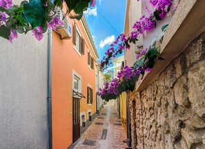 Una calle estrecha flanqueada por edificios, uno pintado de naranja y el otro de un color claro, con un muro de piedra a la derecha.