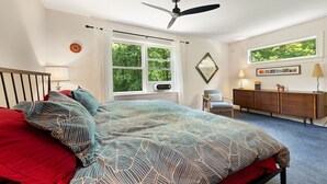 Een slaapkamer met een bed, een plafondventilator, een raam met gordijnen, een stoel, een dressoir en een lamp.