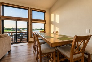 A dining area with a long wooden table, six chairs, and a view through large windows.