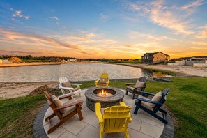 A patio with Adirondack chairs and a fire pit by a lake, with houses in the background. - Sunset Lakefront with Kayaks & Firepit Fun (New Lisbon)