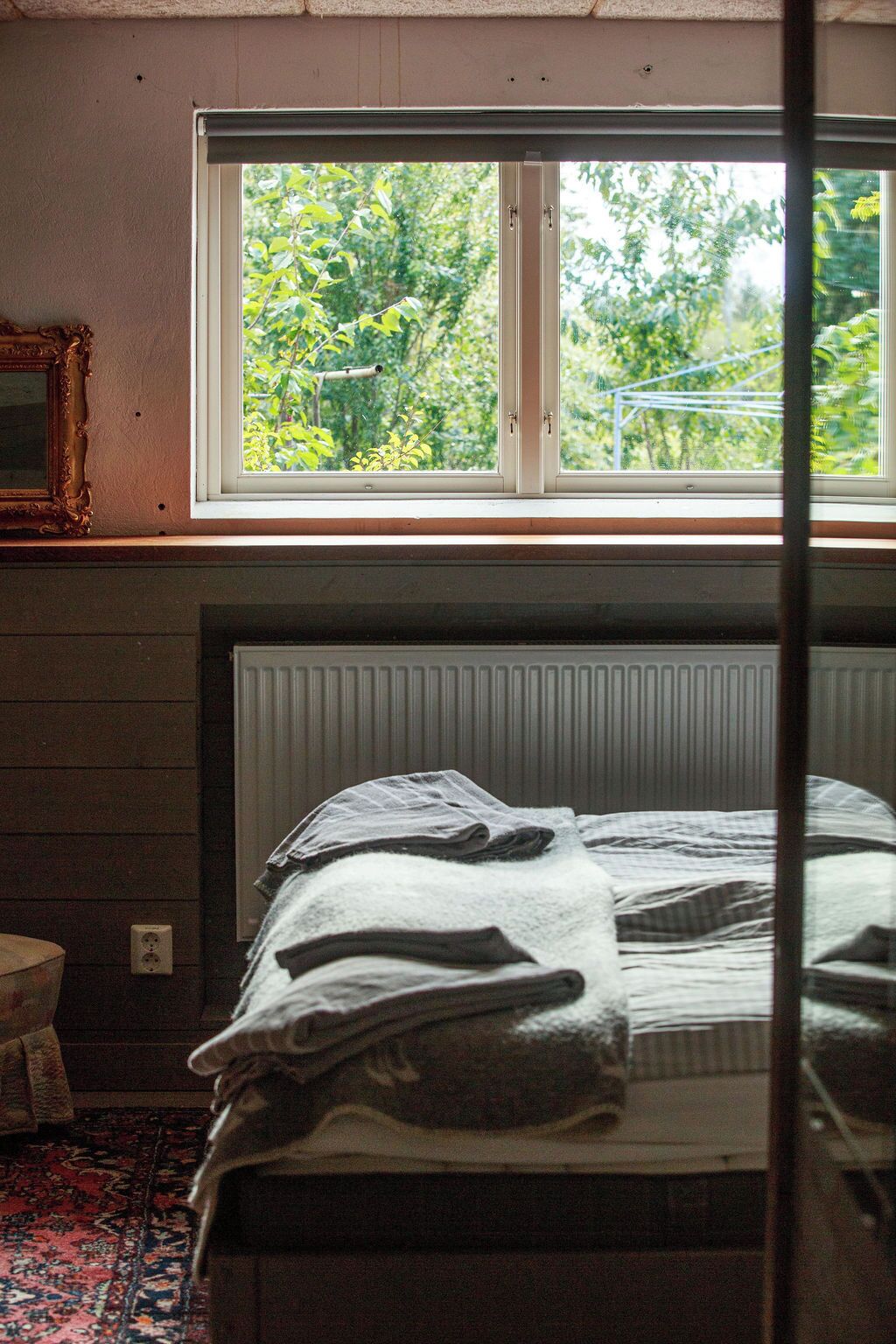A bedroom with a bed, a radiator, a window with a view of greenery, and a mirror on the wall.