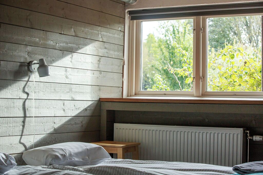 A bedroom with a wooden wall, a window with a view of trees, a radiator, and a bedside table.