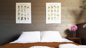Une chambre avec une tête de lit en bois, du linge de lit blanc, un plaid marron et deux affiches à motifs végétaux accrochées au mur.