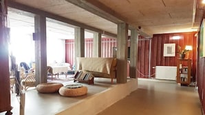 Une pièce spacieuse avec un plafond bas, un canapé recouvert d’une couverture et un radiateur. On y trouve des poufs en osier et une table avec des chaises.