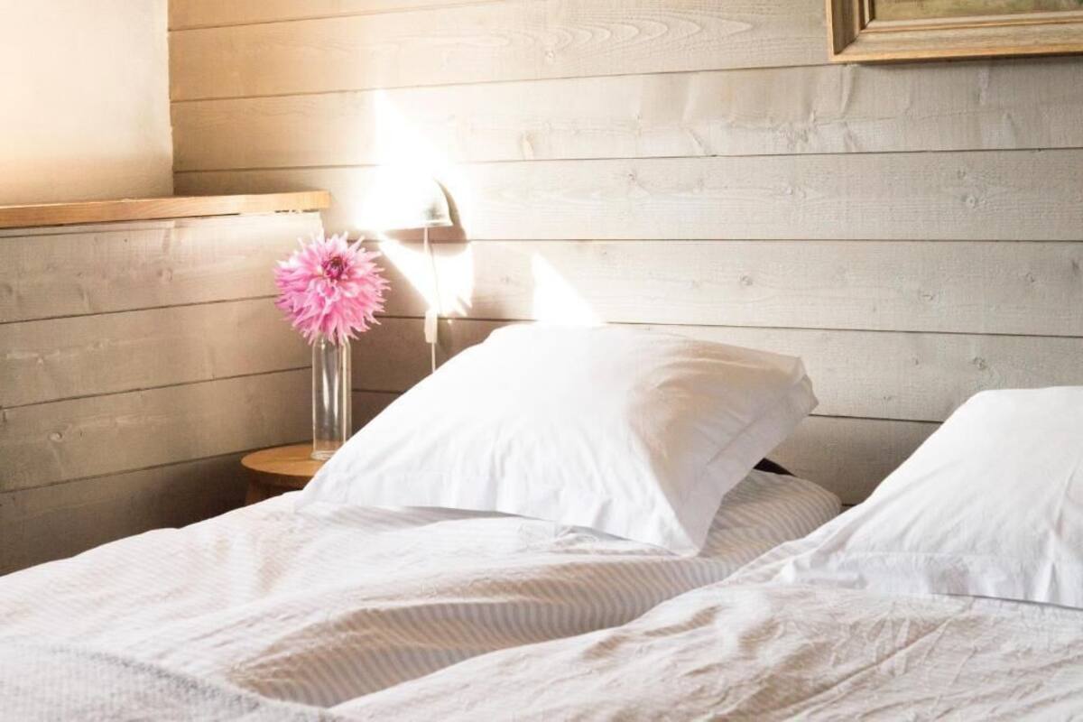 Ein ordentlich bezogenes Bett mit weißen Leinen und Kissen, ein Nachttisch mit einer Vase voller rosa Blumen und ein hölzernes Kopfende.