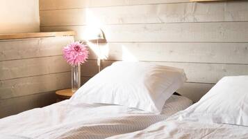 A neatly made bed with white linens and pillows, a bedside table with a vase of pink flowers, and a wooden headboard.