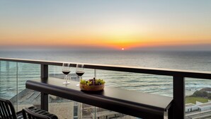 A balcony with a table, two wine glasses, and a bowl of fruit overlooking the ocean at sunset.