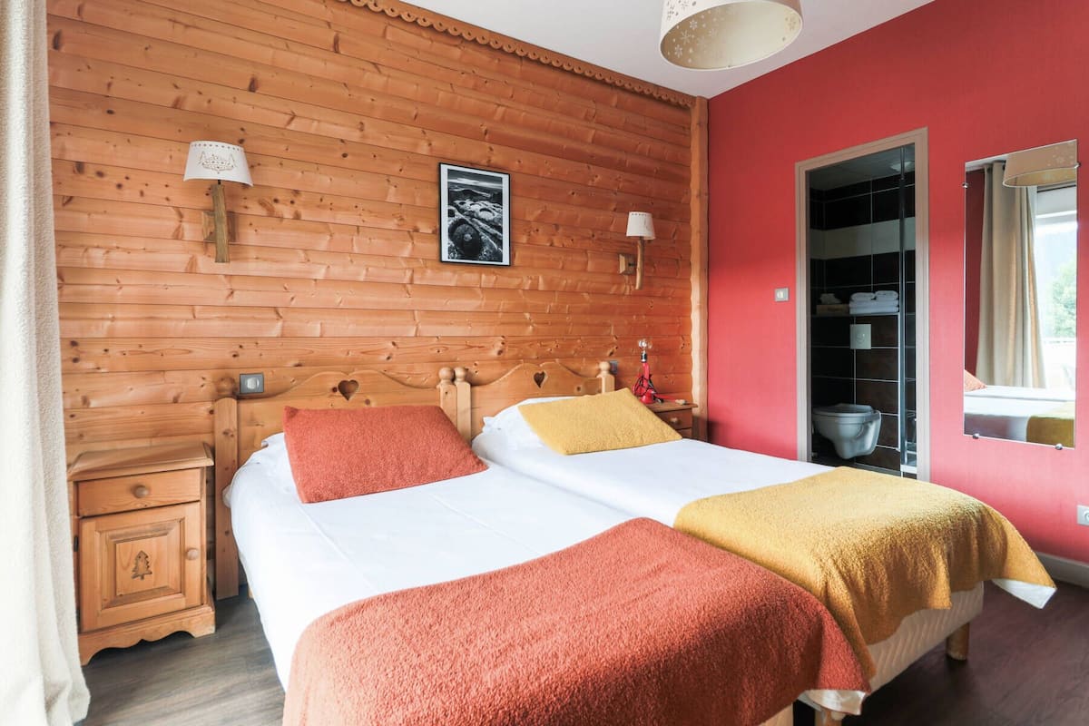 A double bed with two red and one yellow blanket, wooden paneling on the walls, a framed picture, and a window with curtains.