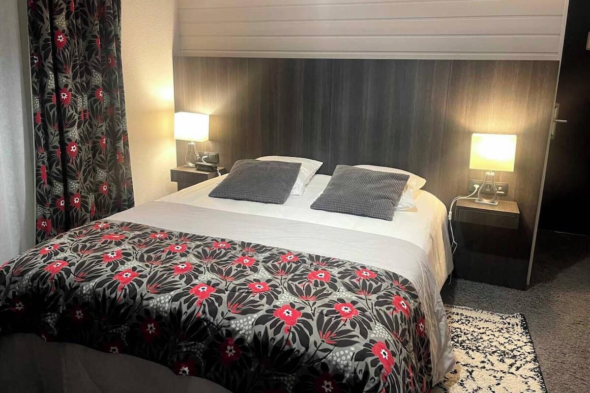 A hotel room with a bed, two bedside tables with lamps, a patterned bedspread, and a window with curtains.