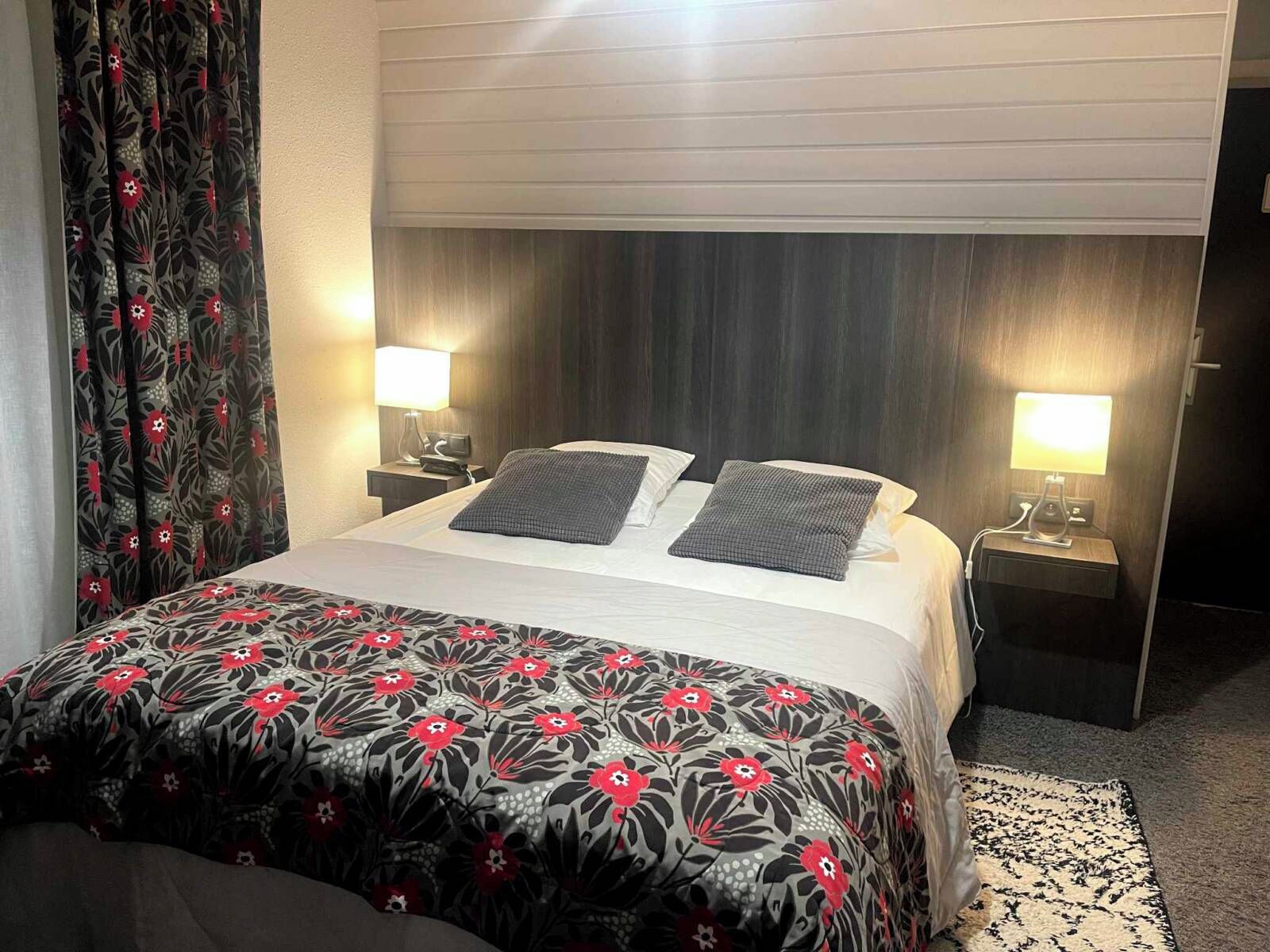 Ein Hotelzimmer mit einem Bett, zwei Nachttischen mit Lampen, einer gemusterten Bettdecke und einem Fenster mit Vorhängen.