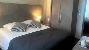 Una habitación de hotel con una cama grande, un armario, una mesita de noche con lámpara y una pequeña mesa con tetera y tazas.