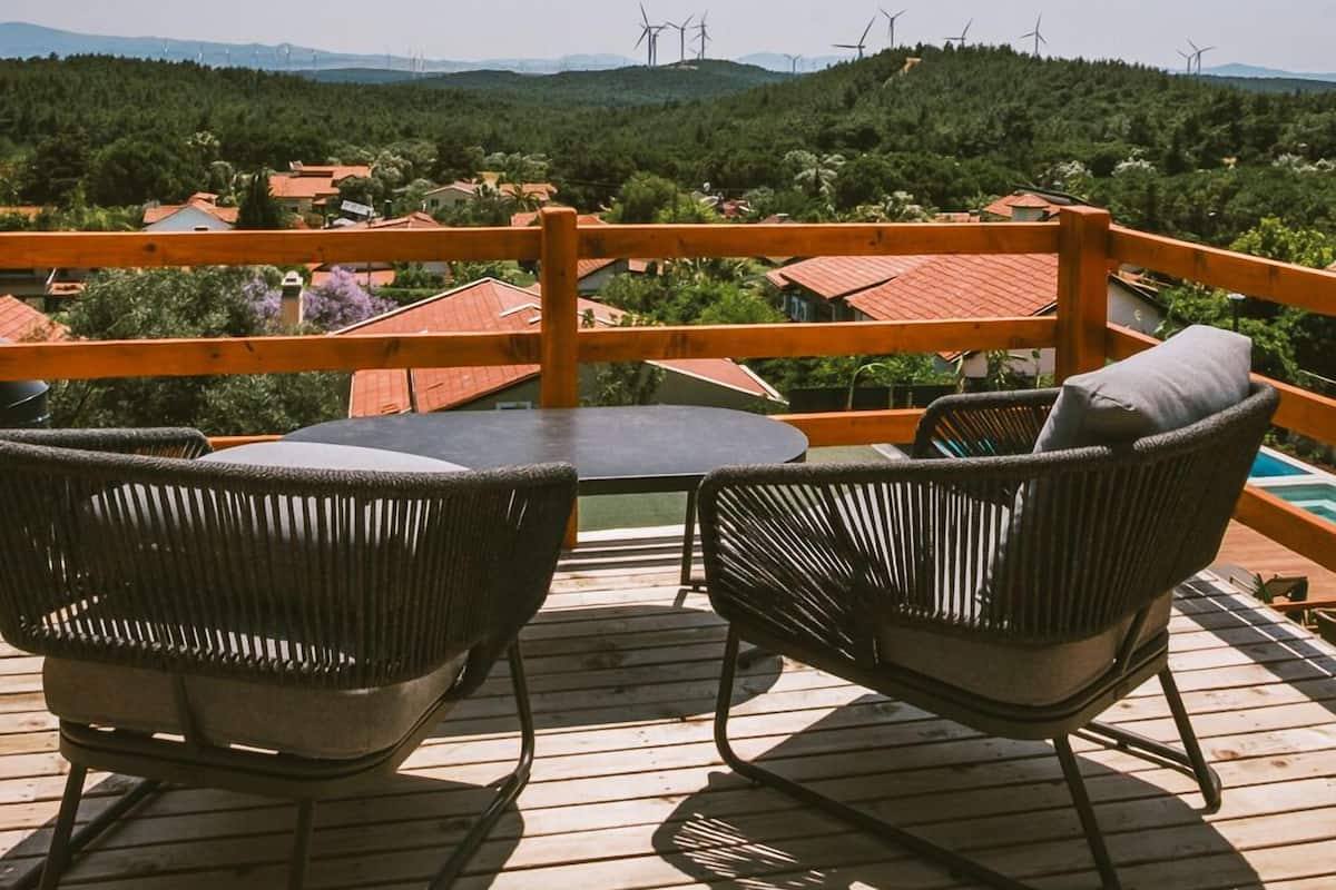 A wooden deck with two wicker chairs and a table, overlooking a village with wind turbines in the distance.
