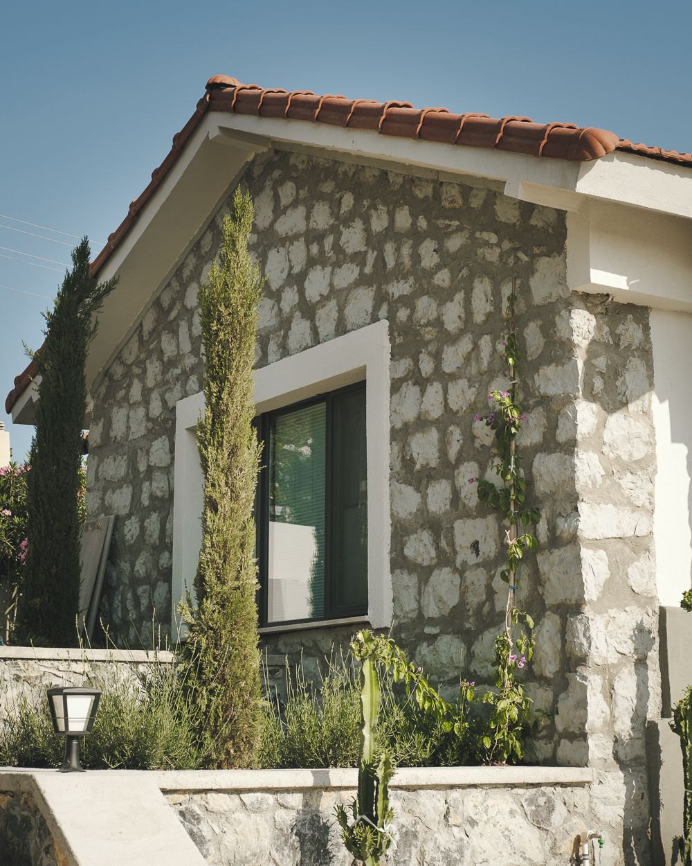 A stone building with a tiled roof, a window, and a small garden area with plants and a lamp post.