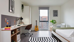 A modern bedroom with a bed, desk, chair, guitar, and a view of buildings through a window.