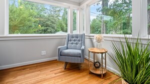 Une pièce dotée de grandes fenêtres, d’un fauteuil à motifs bleus, d’une petite table en bois avec une lampe et d’une plante en pot.