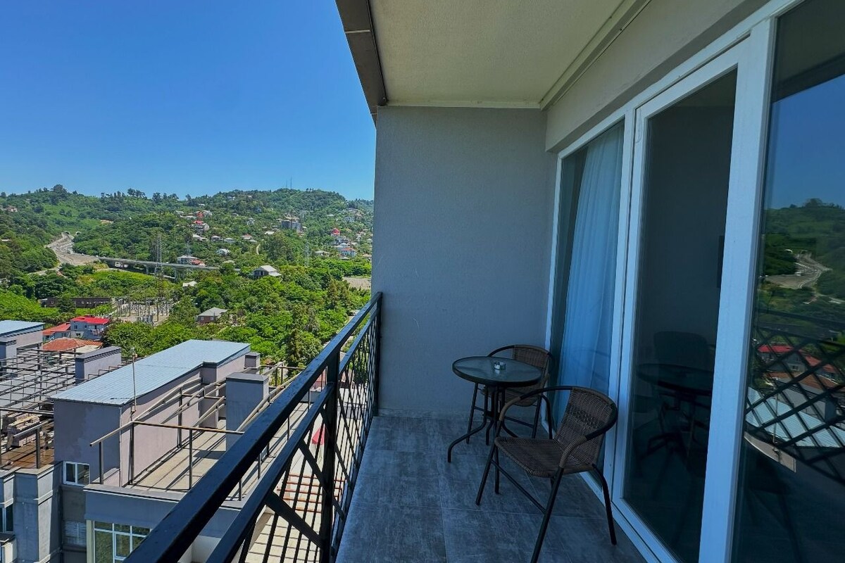 Un balcon avec deux chaises et une table, offrant une vue sur le paysage urbain et la verdure.