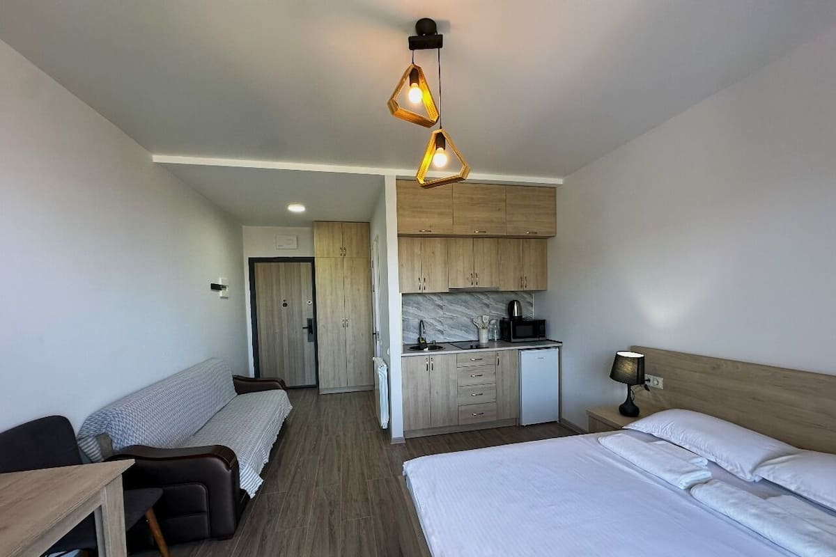 A hotel room with two beds, a kitchenette, a small seating area, and a wooden ceiling light fixture.