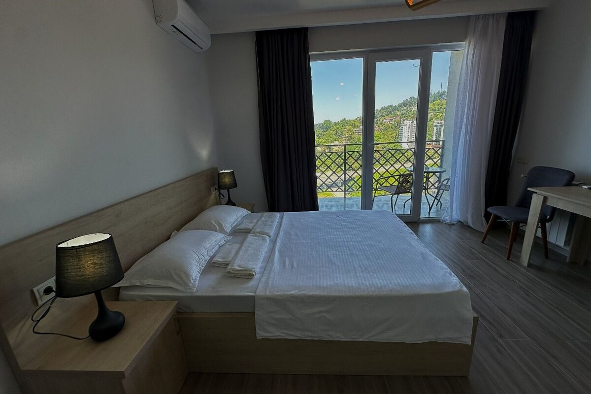 A hotel room with a bed, a desk, a chair, a lamp, and a view of a balcony with a railing.
