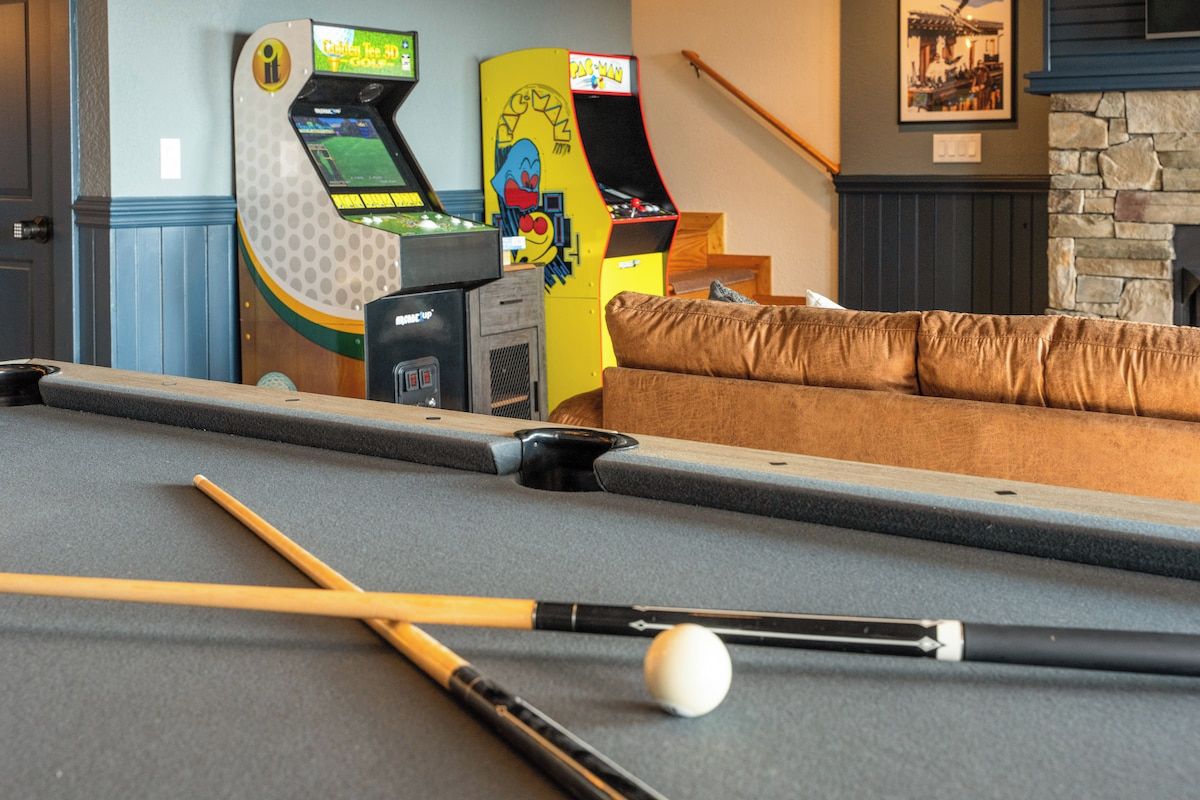 Een pooltafel met poolkeus en een bal, een bank, en twee speelautomaten in een ruimte met een stenen openhaard en een ingelijste afbeelding aan de muur.