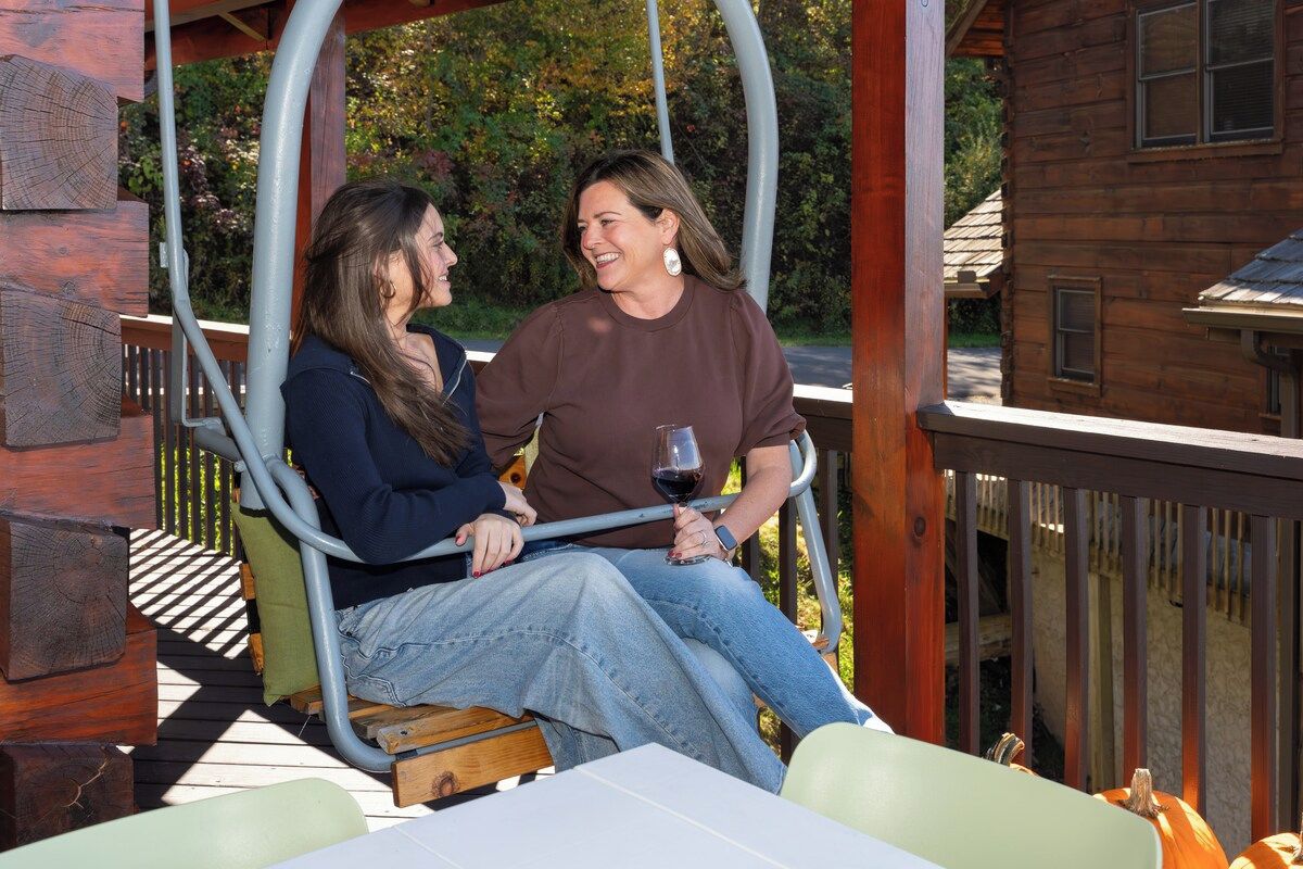 Two individuals seated on a swing chair outdoors, with a wooden structure and a house in the background.
